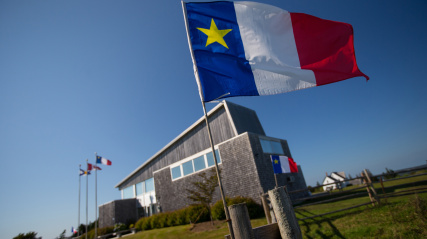 Low-angle shot focusing on a large Acadian flag waving prominently in the foreground on a clear day. The flag is attached to a wooden fence post on a grassy hill. In the background, a modern building with weathered wood-shingled siding sits at a slight angle, with additional flags flying from poles on the far left.