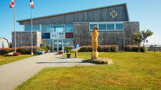 Exterior view of a modern two-story building with weathered wood shingle siding and a sloped roof under a clear blue sky. A paved walkway leads to the glass entrance doors, which feature signs reading "Bienvenue" and "Welcome" alongside the Le Village historique acadien logo. In front of the building, a large green lawn features a wooden statue of a historical figure in a wide-brimmed hat standing on a stone base, flanked by flower beds and two tall flagpoles flying the Canadian flag and the Acadian flag.